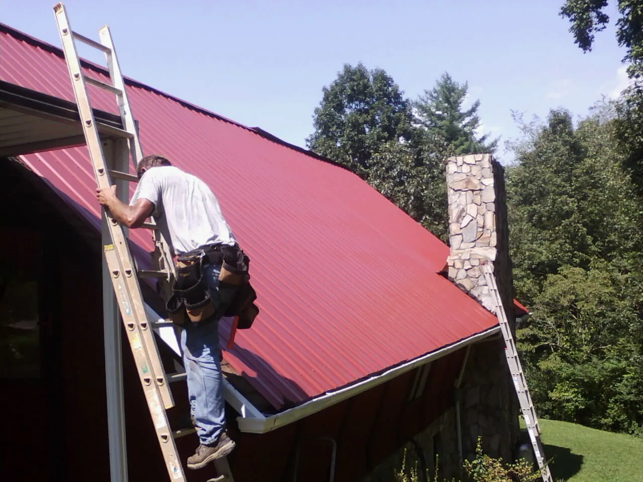 Roofer on a ladder beside a red metal roof and stone chimney — Soffit & Fascia Repair by Roofing Brothers in Claxton